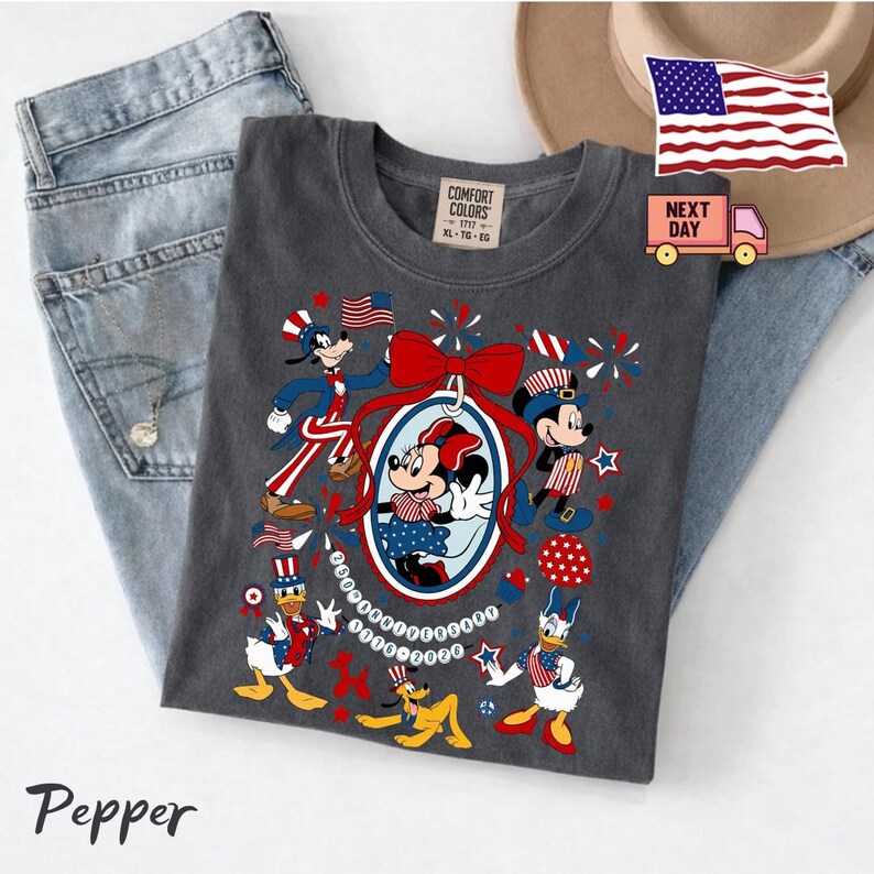 Może przedstawiać: Ciemnoszary t-shirt z grafiką przedstawiającą postacie Disneya, w tym Myszkę Miki, Minnie, Kaczora Donalda, Goofy'ego i Pluto w patriotycznych czerwono-biało-niebieskich strojach. Wz&oacute;r zawiera fajerwerki, gwiazdki oraz napis 100th Anniversary 1928-2028.