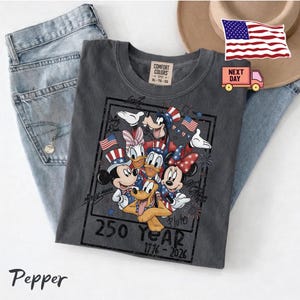 May include: A charcoal gray t-shirt featuring a graphic of Disney characters including Mickey Mouse, Minnie Mouse, Donald Duck, Daisy Duck, Goofy, and Pluto wearing patriotic attire. The text reads 250 Year 1776-2026 with small American flags.