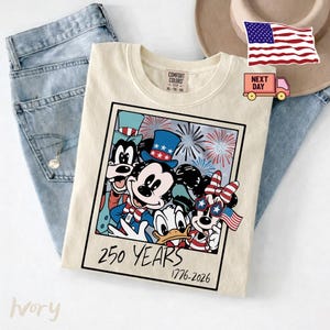 May include: An ivory t-shirt featuring a graphic print of Mickey Mouse, Minnie Mouse, Donald Duck, and Goofy wearing patriotic hats and accessories. The design includes fireworks and the text 250 YEARS 1776-2026 in a square frame.