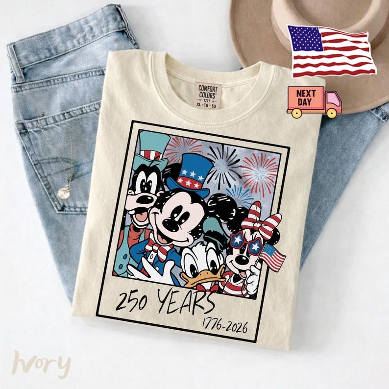 May include: An ivory t-shirt featuring a graphic print of Mickey Mouse, Minnie Mouse, Donald Duck, and Goofy wearing patriotic hats and accessories. The design includes fireworks and the text 250 YEARS 1776-2026 in a square frame.