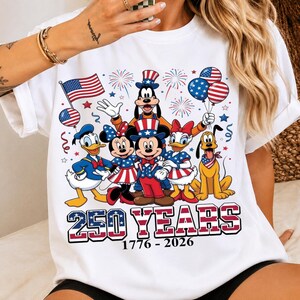 Może przedstawiać: Biały t-shirt z grafiką przedstawiającą postacie Disneya, w tym Myszkę Miki, Myszkę Minnie, Kaczora Donalda, Daisy, Goofy'ego i Pluto w patriotycznych strojach w kolorach czerwonym, białym i niebieskim. Napis głosi 250 YEARS 1776 - 2026 z fajerwerkami.