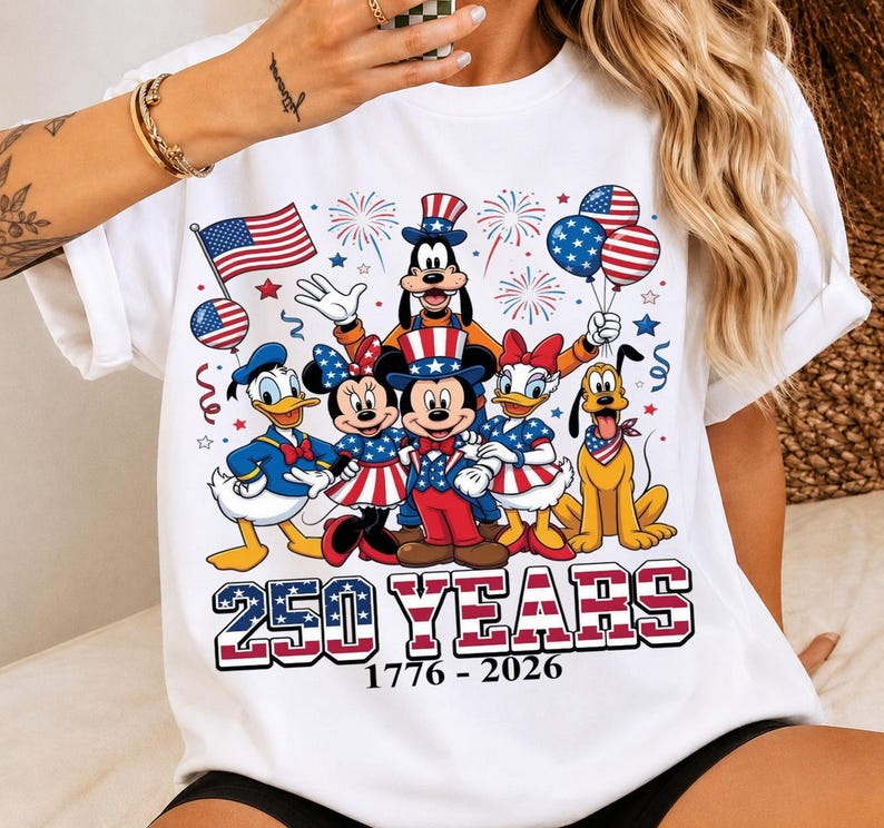 Może przedstawiać: Biały t-shirt z grafiką przedstawiającą postacie Disneya, w tym Myszkę Miki, Myszkę Minnie, Kaczora Donalda, Daisy, Goofy'ego i Pluto w patriotycznych strojach w kolorach czerwonym, białym i niebieskim. Napis głosi 250 YEARS 1776 - 2026 z fajerwerkami.
