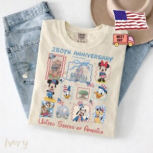 Peut inclure: Un t-shirt ivoire arborant un graphique 250th Anniversary 1776-2024 avec des illustrations de Mickey, Minnie, Donald, Dingo, Pluto, Tic, Tac et Daisy, ainsi que des monuments et le texte United States of America.