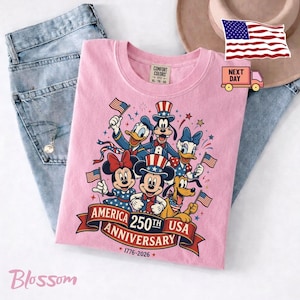 May include: A light pink t-shirt featuring a graphic of Mickey Mouse, Minnie Mouse, Donald Duck, Daisy Duck, Goofy, and Pluto wearing patriotic hats. The text reads AMERICA 250TH ANNIVERSARY USA 1776-2026. The shirt is displayed with blue jeans.