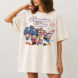 May include: An off-white t-shirt featuring a graphic print of cartoon characters in patriotic attire. The text America Vibes is printed above a map of the United States filled with stars and stripes, accompanied by fireworks illustrations.