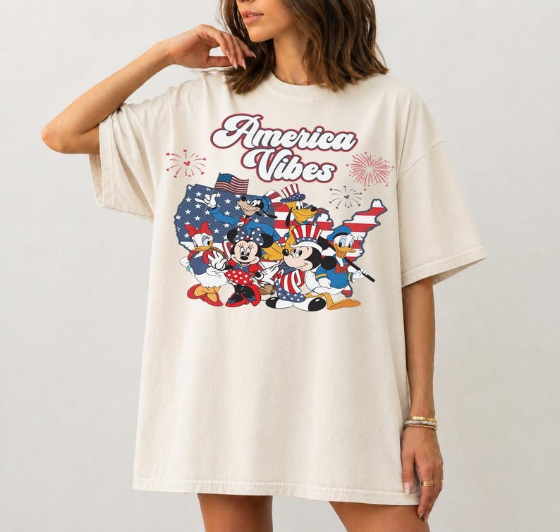 May include: An off-white t-shirt featuring a graphic print of cartoon characters in patriotic attire. The text America Vibes is printed above a map of the United States filled with stars and stripes, accompanied by fireworks illustrations.