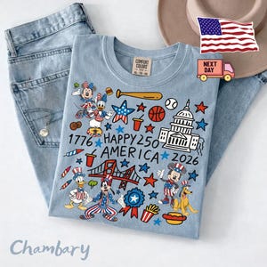 May include: A light blue t-shirt featuring a graphic print with Disney characters, the US Capitol building, a bridge, and patriotic symbols. The text reads 1776 HAPPY 250 AMERICA 2026. The shirt is displayed alongside light blue denim jeans.