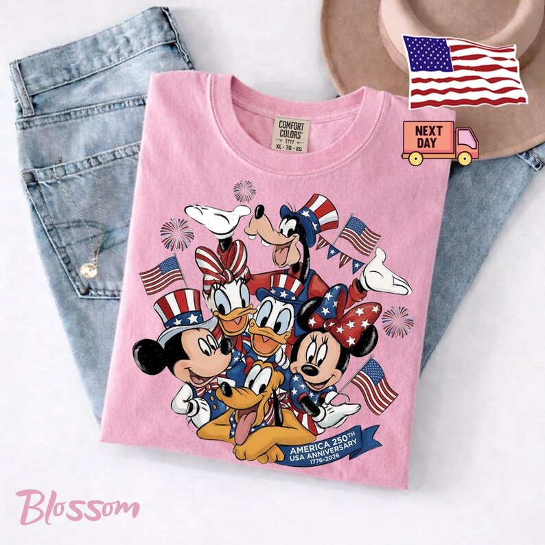 May include: A light pink t-shirt featuring a graphic of Disney characters wearing patriotic hats and outfits, surrounded by fireworks and American flags. The text reads America 250th USA Anniversary 1776-2026. The shirt has a Comfort Colors label.