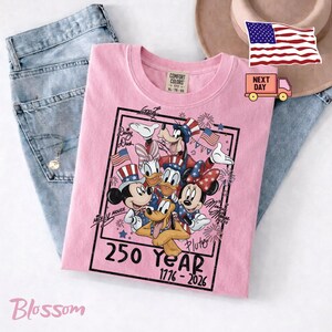 May include: A light pink t-shirt featuring a graphic of Mickey Mouse, Minnie Mouse, Donald Duck, Daisy Duck, Goofy, and Pluto wearing patriotic hats. The text reads 250 YEAR 1776 - 2026, with character names written in script around the black rectangular frame.