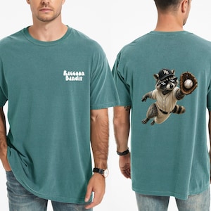 Può includere: Una t-shirt a maniche corte color verde acqua con la scritta bianca Raccoon Bandit sul petto a sinistra. Il retro presenta la grafica di un procione con cappellino da baseball e occhiali pixelati che afferra una palla con un guanto.