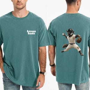 Può includere: Una t-shirt a maniche corte color verde acqua con la scritta bianca Raccoon Bandit sul petto a sinistra. Il retro della maglietta presenta la grafica di un procione che indossa un'uniforme da baseball, un berretto e un guantone da lancio.