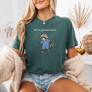 May include: A dark green t-shirt featuring a graphic of a raccoon wearing blue medical scrubs and riding a skateboard. Above the raccoon, the text reads "Off to prevent falls" in a white, simple font. The shirt has a crew neckline and short sleeves.