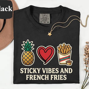 Puede incluir: Una camiseta negra de cuello redondo con un gráfico de una piña, un corazón rojo y una porción de patatas fritas con una pequeña salsa. Debajo del gráfico, el texto dice STICKY VIBES AND FRENCH FRIES en una fuente sans-serif de color blanco roto.