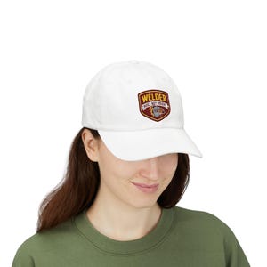 May include: A white baseball cap featuring a shield-shaped patch on the front. The patch has a dark red border and contains the text "WELDER" in yellow letters above the text "BUILT NOT BOUGHT" in white, with a small welding mask graphic below.