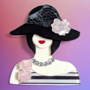 May include: A brooch shaped like a stylized figure wearing a wide-brimmed black hat with a lace overlay and a pale pink flower. The figure has black hair, red lips, a clear rhinestone necklace, and a striped top with two pale pink floral accents.