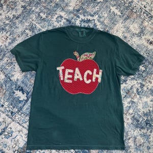 May include: A dark teal short-sleeved t-shirt featuring a large red apple appliqué with white polka dots. The word TEACH is stitched across the centre of the apple in a light-coloured floral fabric. A small patterned leaf is attached to the apple stem.