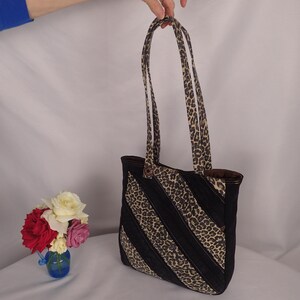 Pode incluir: Uma bolsa tote com um padrão de riscas diagonais de tecido preto e tecido com estampado de leopardo. A bolsa tem duas alças de ombro a condizer com estampado de leopardo e um forro interior castanho escuro visível na abertura superior.