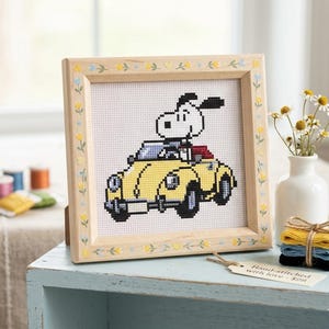Peut inclure: Image d'une broderie au point de croix représentant un chien de dessin animé dans une décapotable jaune. Le cadre en bois clair est orné de motifs floraux. Un petit vase de fleurs et des bobines de fil sont à proximité. L'œuvre est brodée à la main.