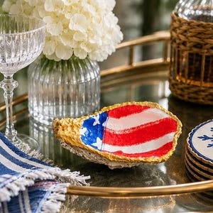 May include: An oyster shell painted with the American flag design, featuring red and white stripes and a blue field with white stars. The shell has a gold-colored edge and sits on a reflective surface. A glass of water and a vase of white flowers are in the background.
