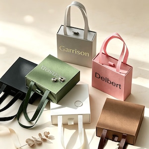 Può includere: Una collezione di sei scatole regalo quadrate con manici, nei colori nero, verde oliva, grigio chiaro, bianco, rosa e marrone. La scatola grigia riporta la scritta Garrison, quella rosa Delbert e quella verde oliva la scritta Benson.