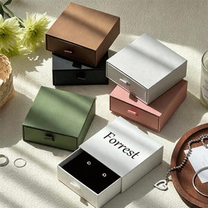 Può includere: Una collezione di scatole quadrate a cassetto in marrone, nero, verde oliva, bianco, grigio chiaro e rosa polvere. La scatola bianca presenta la parola Forrest stampata in nero sul coperchio, con un interno nero che contiene due piccoli orecchini.