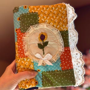 May include: A quilted fabric book cover featuring a patchwork design of yellow, green, orange, and blue floral patterns. The center displays an embroidered sunflower inside a lace-trimmed circular frame with a small white ribbon bow and white eyelet lace trim.