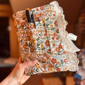 May include: Floral print quilted notebook cover with pockets, featuring orange, yellow, and white flowers on a cream background. The cover has a white lace trim and a white ribbon closure. Pens are visible in the pockets.