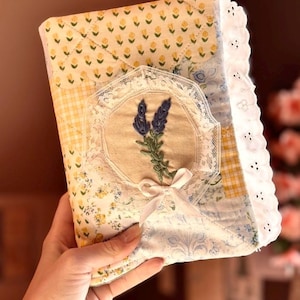 May include: A quilted fabric book cover featuring a patchwork design of yellow floral, blue patterned, and yellow gingham fabrics. The center displays an embroidered lavender sprig framed by white lace and a small white ribbon bow, with white eyelet trim.