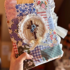 May include: A quilted book cover with a patchwork design in various floral and patterned fabrics. The cover features a central embroidered cross with blue and brown thread, surrounded by lace trim and a cream-colored bow.
