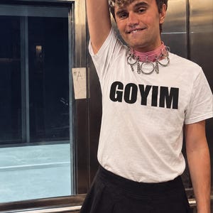 May include: A white short-sleeved t-shirt with the word GOYIM printed in bold black capital letters across the chest, paired with a light pink choker necklace featuring multiple silver-toned metal rings and a black pleated mini skirt.