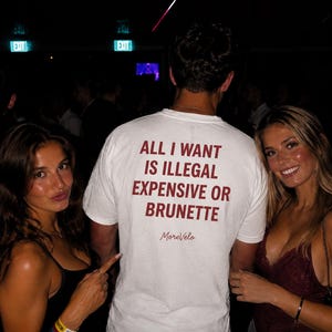May include: A white short-sleeved t-shirt featuring dark red text on the back that reads ALL I WANT IS ILLEGAL EXPENSIVE OR BRUNETTE with the script text MoreVelo underneath. The shirt is worn by a person facing away from the camera.