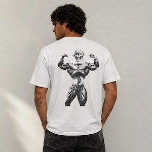 May include: A white short-sleeved t-shirt featuring a black and white graphic on the back. The graphic depicts a muscular figure with a skull head, flexing its arms in a pose. The figure has a detailed, textured appearance with visible muscle definition.
