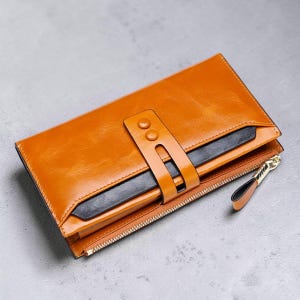 May include: A rectangular mustard yellow wallet featuring a smooth finish, a central vertical strap secured with two snap buttons, a contrasting black horizontal accent panel, and a side zip closure with a matching mustard yellow pull tab.