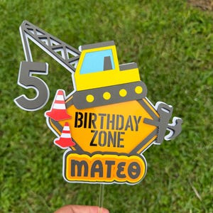 May include: A construction-themed cake topper featuring a yellow excavator, a gray crane arm, and the number 5. It includes a yellow hexagonal sign with the text BIRTHDAY ZONE, two red and white traffic cones, a saw, a hammer, and the name MATEO.
