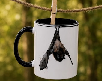 May include: A white ceramic mug with a black handle and interior rim, featuring a watercolor-style illustration of a brown and black bat hanging upside down. The mug is suspended from a brown rope by a wooden clothespin.