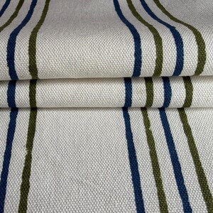 May include: A woven fabric featuring a repeating pattern of vertical stripes in navy blue and olive green against an off-white background. The textile has a visible textured weave and is folded to show depth and the consistent striped design.