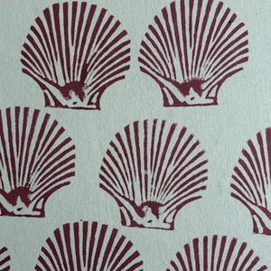 May include: A light mint green fabric featuring a repeating block print pattern of dark maroon scallop shells. The shells are arranged in a grid-like formation, each displaying detailed radiating lines and a textured, hand-stamped appearance.