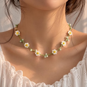 May include: A delicate necklace featuring a thin green cord adorned with small, crocheted white daisy flowers with bright yellow centers. The floral design is spaced evenly along the length of the cord, creating a simple, nature-inspired accessory.