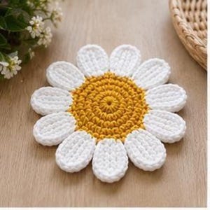 May include: A round, crocheted coaster shaped like a daisy flower. The center is a textured, circular mustard yellow yarn pattern, surrounded by twelve individual white, oval-shaped petals made of thick, looped yarn stitches.