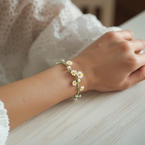 Könnte beinhalten: Ein zartes Armband mit einem dünnen grünen Fadenband, das mit kleinen, gehäkelten weißen Gänseblümchen mit gelben Mitten verziert ist. Das florale Design schlingt sich um das Handgelenk und zeigt eine filigrane Handarbeit.