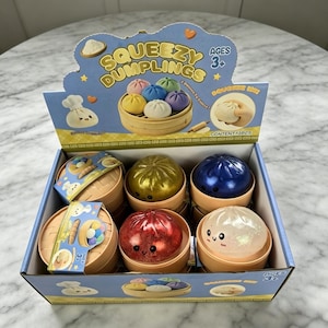 Op de afbeelding: Een winkeldisplaydoos met het label Squeezy Dumplings met twaalf individuele speelgoedknoedels. Elke knoedel zit in een klein, rond, beige plastic stoommandje. De set bevat metallic gouden, blauwe, rode en doorzichtige knoedelspeeltjes.