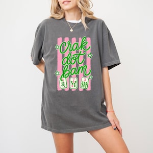 May include: A charcoal grey short-sleeve t-shirt featuring a graphic with vertical pink stripes and the green text "crak dot bam". Below the text are three mahjong tiles showing the numbers 5, 7, and 3 with green and pink details.