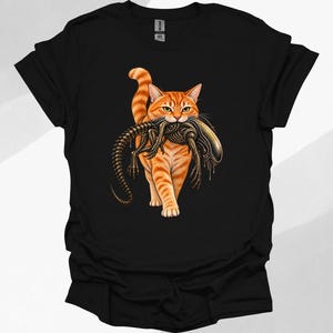 Peut inclure: Un t-shirt noir à manches courtes avec une impression graphique représentant un chat tigré orange marchant vers l'avant tout en portant une créature sombre, squelettique et extraterrestre dans sa gueule aux yeux verts.