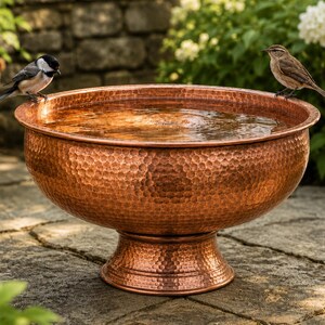 May include: A round, hammered copper birdbath with a wide basin and a pedestal base, filled with water. A black and white chickadee sits on the left rim, while a brown and tan bird rests on the right rim of the metallic, textured vessel.
