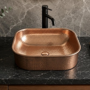 May include: A square vessel sink with a hammered copper finish and a central drain. The sink is paired with a tall, matte black faucet featuring a single lever handle. The basin has rounded corners and a reflective, textured metallic surface.