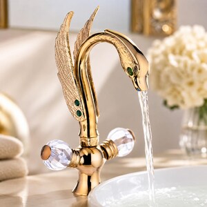 May include: A gold-toned swan-shaped faucet with textured wings, green gemstone accents, and two clear, faceted crystal handles. Water flows from the swan's beak into a white basin, highlighting the metallic finish and intricate detailing.
