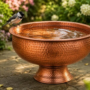 May include: A round, hammered copper birdbath with a wide basin and a pedestal base, filled with clear water. A small black, white, and gray chickadee perches on the rim of the basin, looking down into the water. The metal surface has a textured finish.