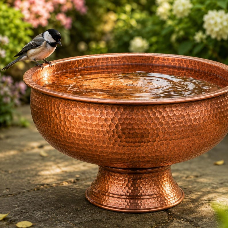 May include: A round, hammered copper birdbath with a wide basin and a pedestal base, filled with clear water. A small black, white, and gray chickadee perches on the rim of the basin, looking down into the water. The metal surface has a textured finish.