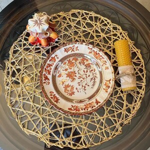 May include: A table setting with a woven gold placemat, a plate with floral patterns, a candle wrapped in burlap, and a star-shaped candle. The plate has a white background with orange and brown floral designs.