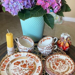 May include: A table setting with floral china plates and teacups, featuring orange and gold detailing. A bouquet of purple and pink hydrangeas is in a turquoise vase. A yellow candle and a decorative candle are also present.